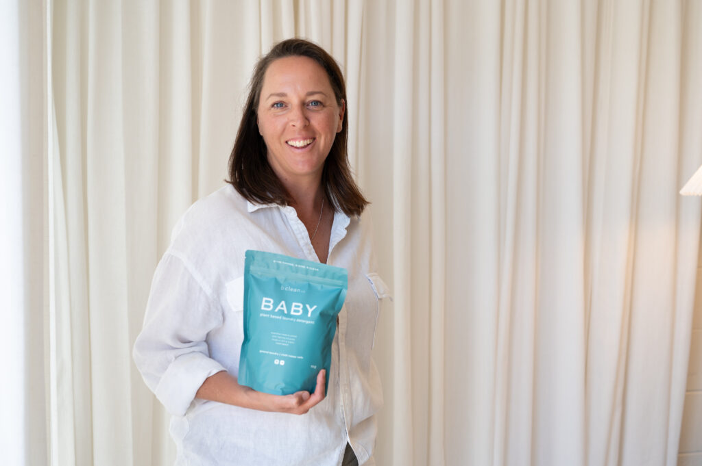 leanne in white shirt holding baby detergent with cream background curtain.