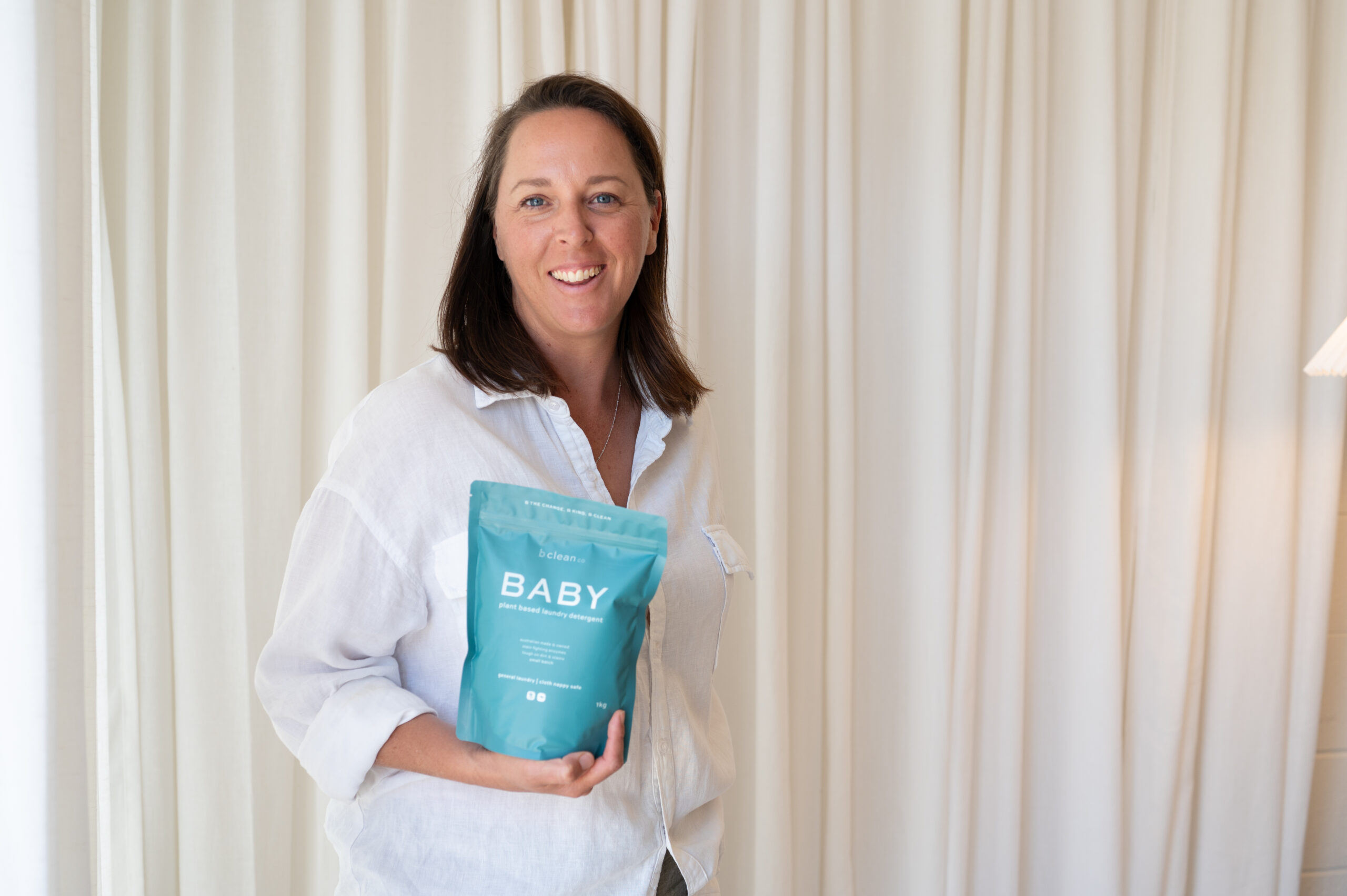 leanne in white shirt holding baby detergent with cream background curtain.